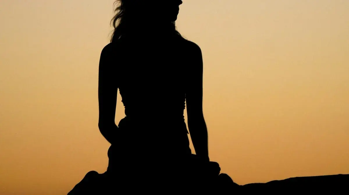 A woman seated in silhouette against a warm amber sunset sky, body still and contemplative. The embodied quiet The Body Knows movement guide is built around.